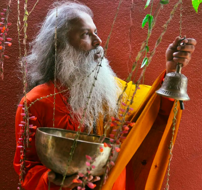 AkilLanka con túnica naranja sosteniendo un cuenco tibetano y una campana ritual frente a un fondo rojo